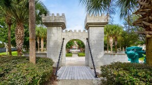 A stone archway with a wooden pathway leading through it, surrounded by palm trees and greenery. - Windsor Hills Resort Homes by Atlas VH (Kissimmee)