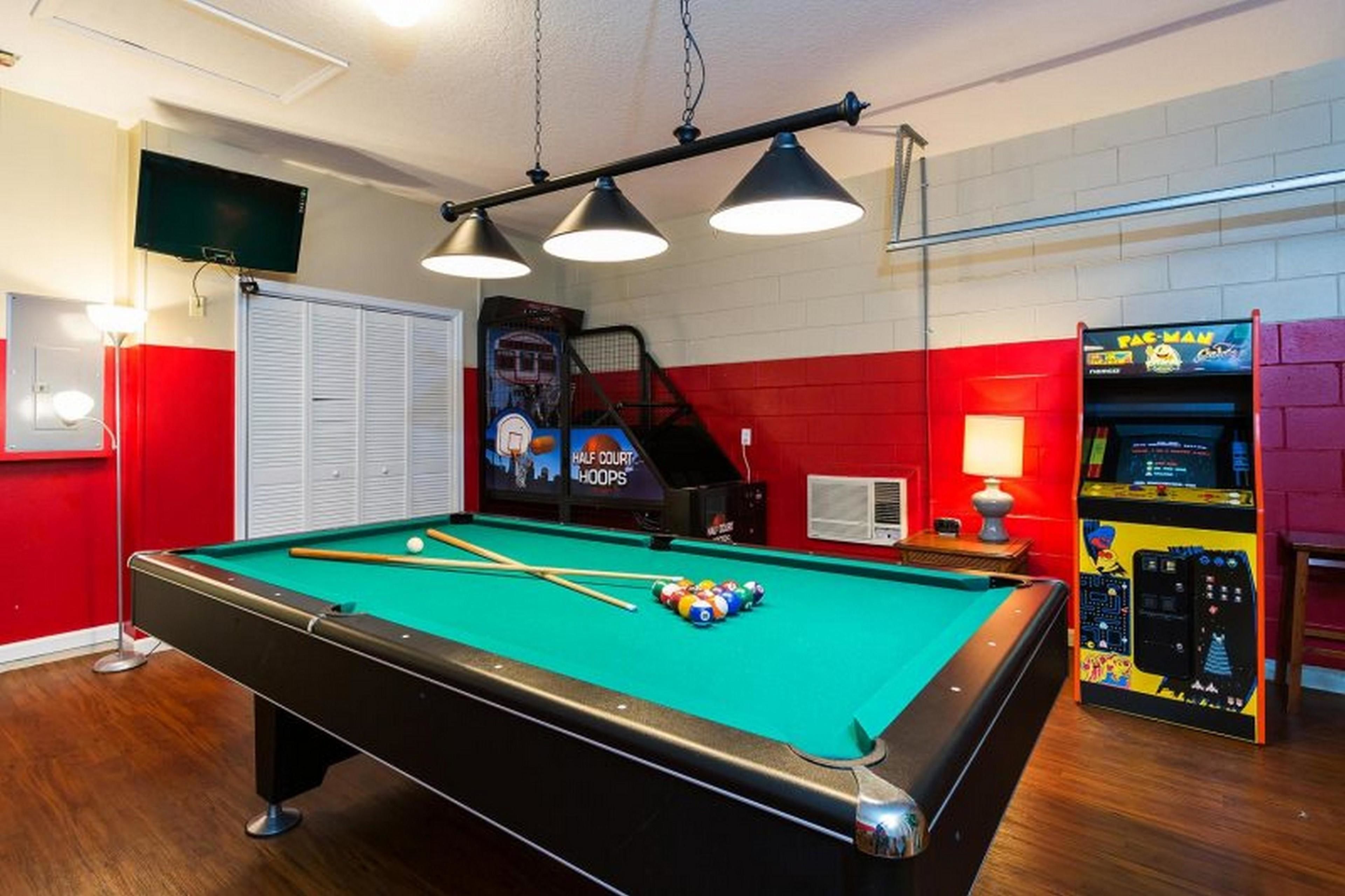 Een pooltafel met drie hanglampen, een televisie en een Pac-Man spelcomputer in een kamer met rode en witte wanden.