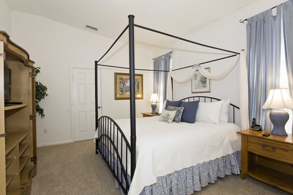 A four-poster canopy bed with a white and blue floral bedspread, a wooden nightstand with a lamp, and a door with a framed picture.