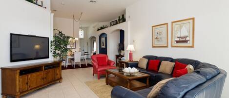 A living room with a television, sofa, red chair, coffee table, and a dining area in the background.