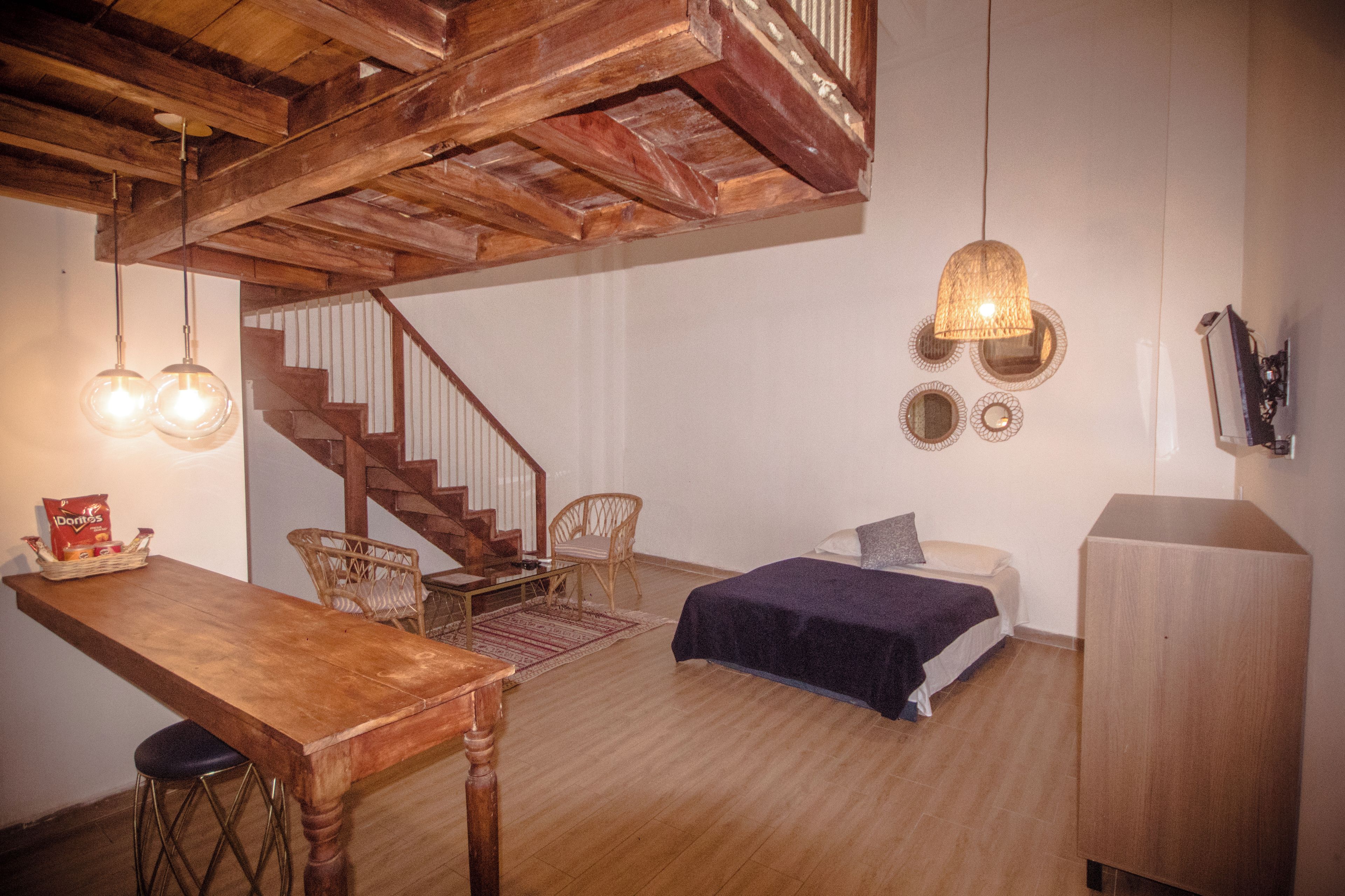 Una habitación con una escalera de madera, una cama con una manta azul, una mesa de madera con sillas y un televisor montado en la pared.