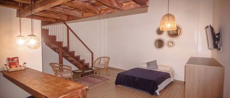 Una habitación con una escalera de madera, una cama con una manta azul, una mesa de madera con sillas y un televisor montado en la pared.