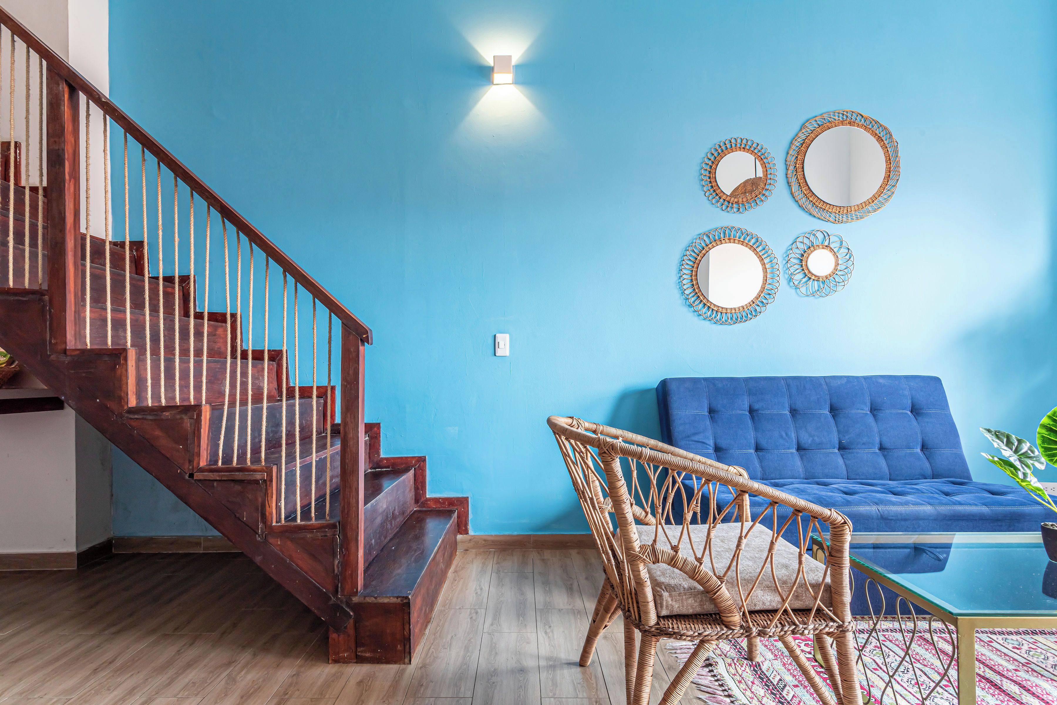 A living room with a blue sofa, a wicker chair, a glass coffee table, and a wooden staircase.