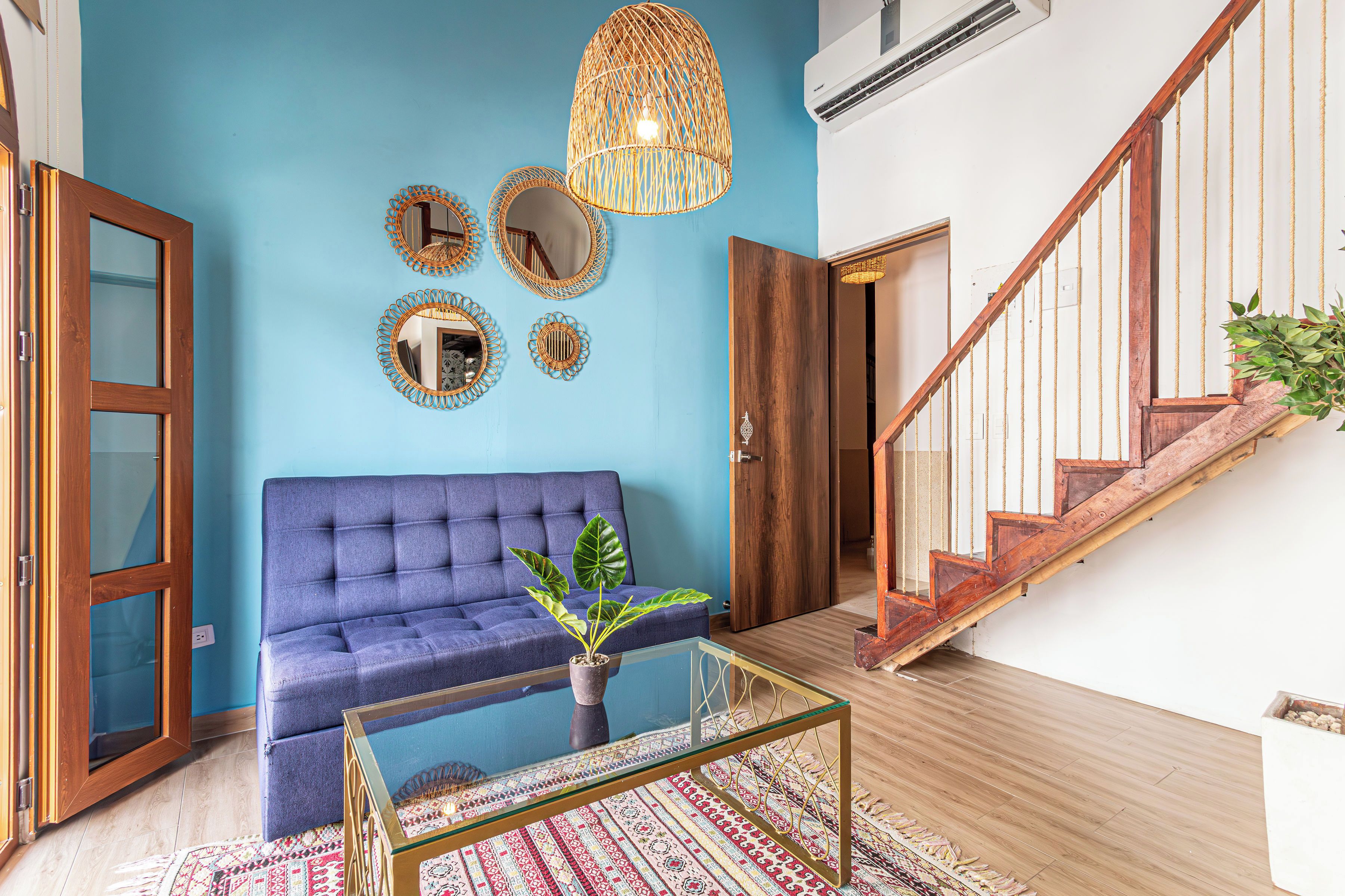 Una habitación con un sofá azul, una mesa de centro de vidrio, una alfombra estampada, una escalera de madera y una pared con espejos decorativos.