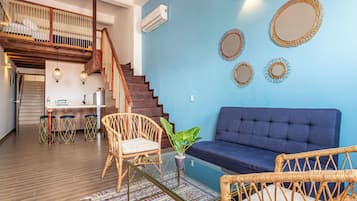 Een woonkamer met een blauwe bank, rieten stoelen, een bijzettafeltje en een trap met een houten leuning.