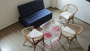 Un salón con un sofá azul oscuro, dos sillones de mimbre con cojines, una mesa de centro de cristal y una alfombra estampada.