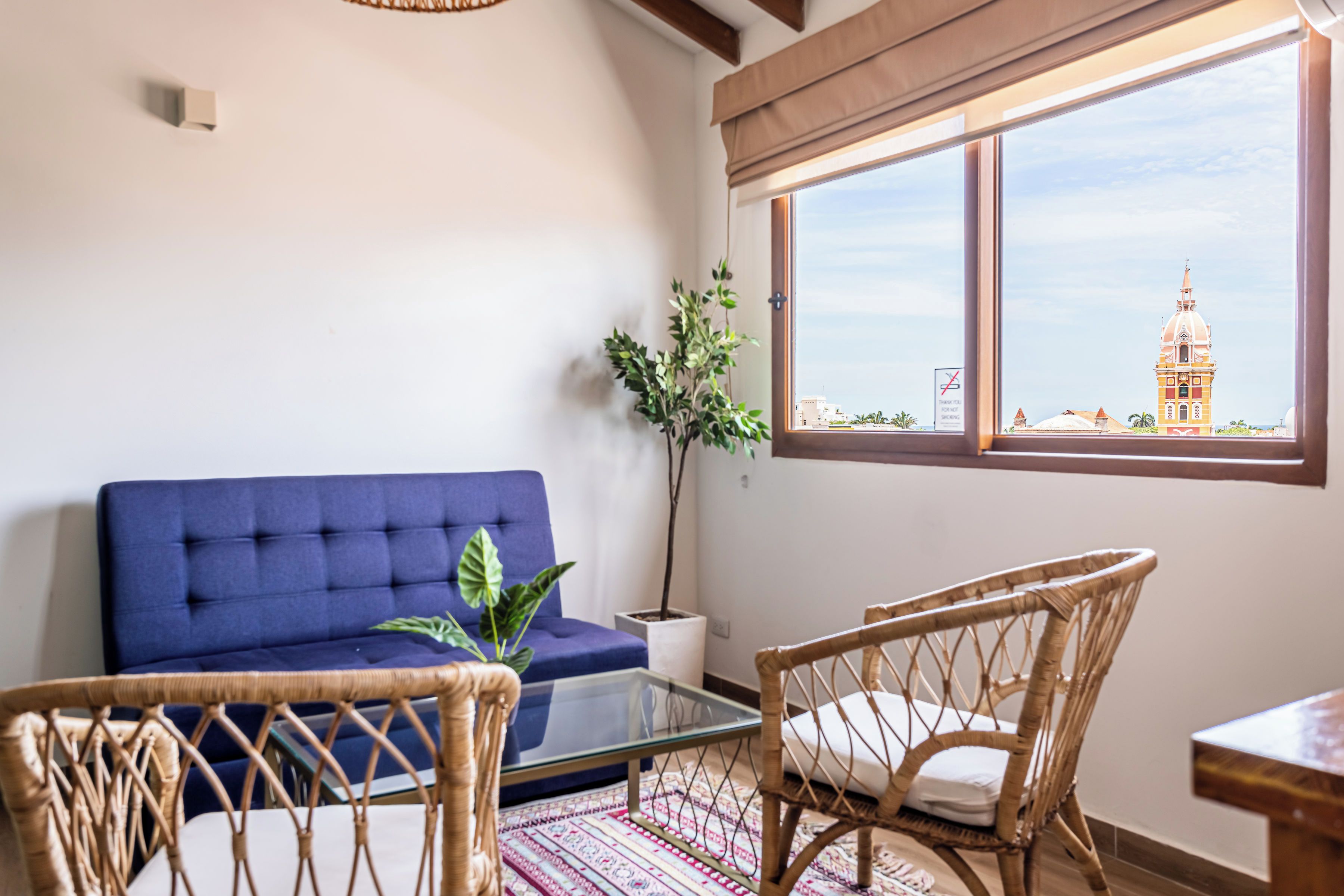 A living room with a blue sofa, a glass coffee table, wicker chairs, a potted plant, and a view of a building through a window.