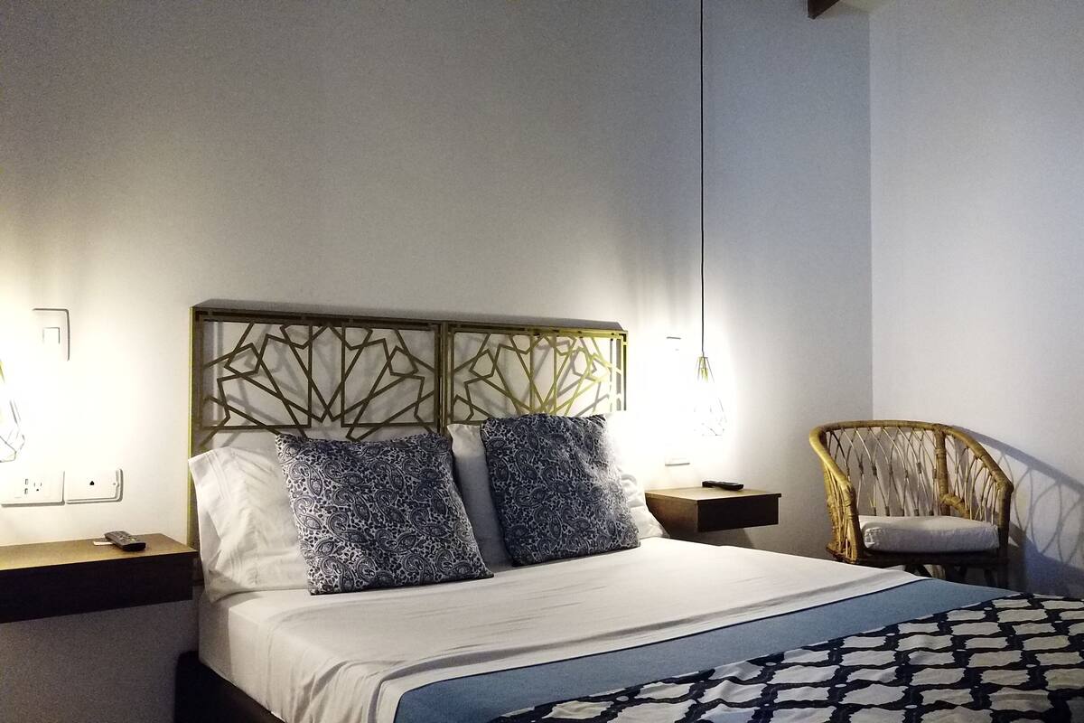 A neatly made bed with patterned pillows, a headboard with geometric designs, a bedside lamp, and a wicker chair.