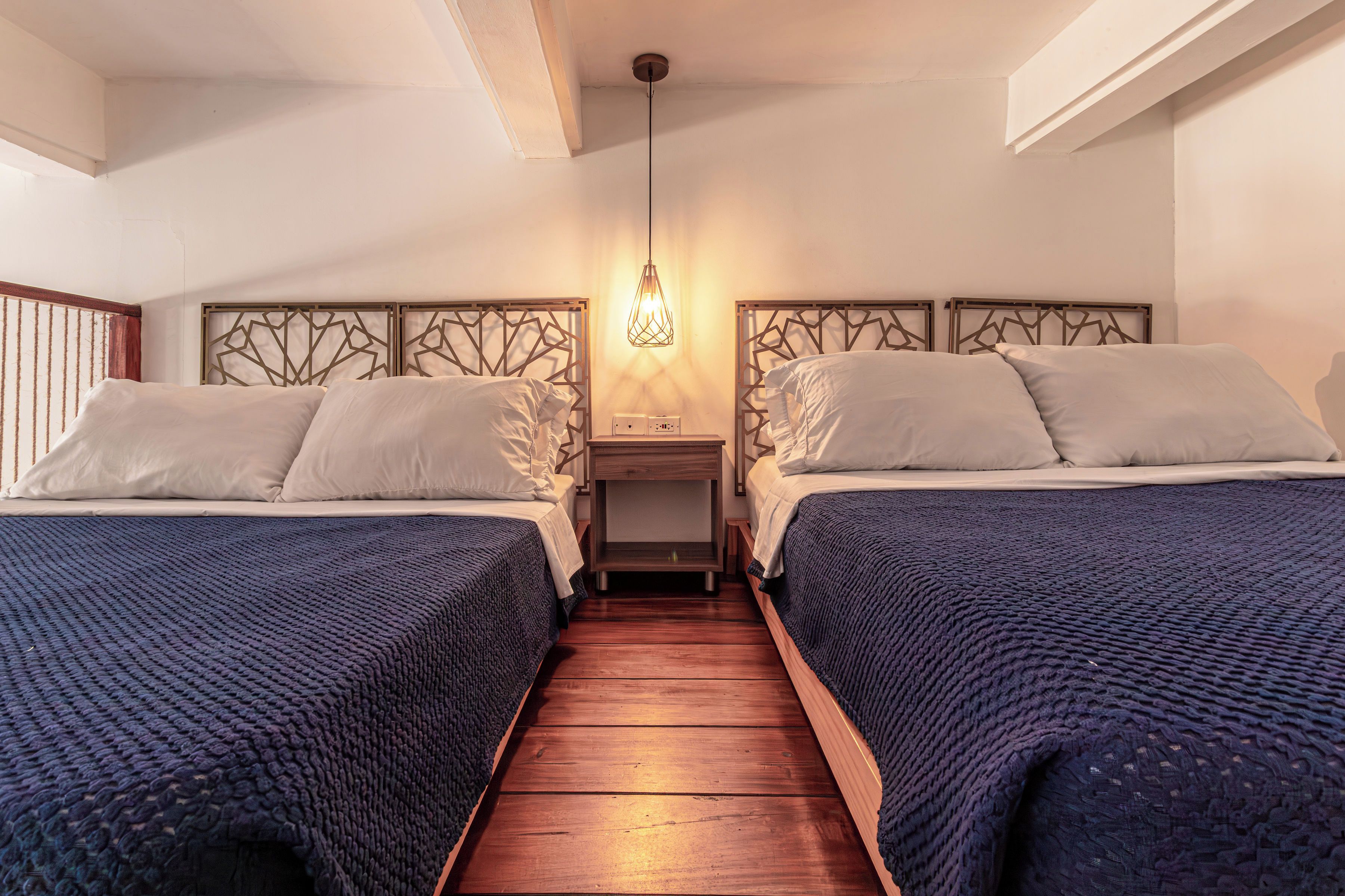 Quarto de hotel com duas camas, piso de madeira, mesa de cabeceira com abajur e painel de cama decorativo.