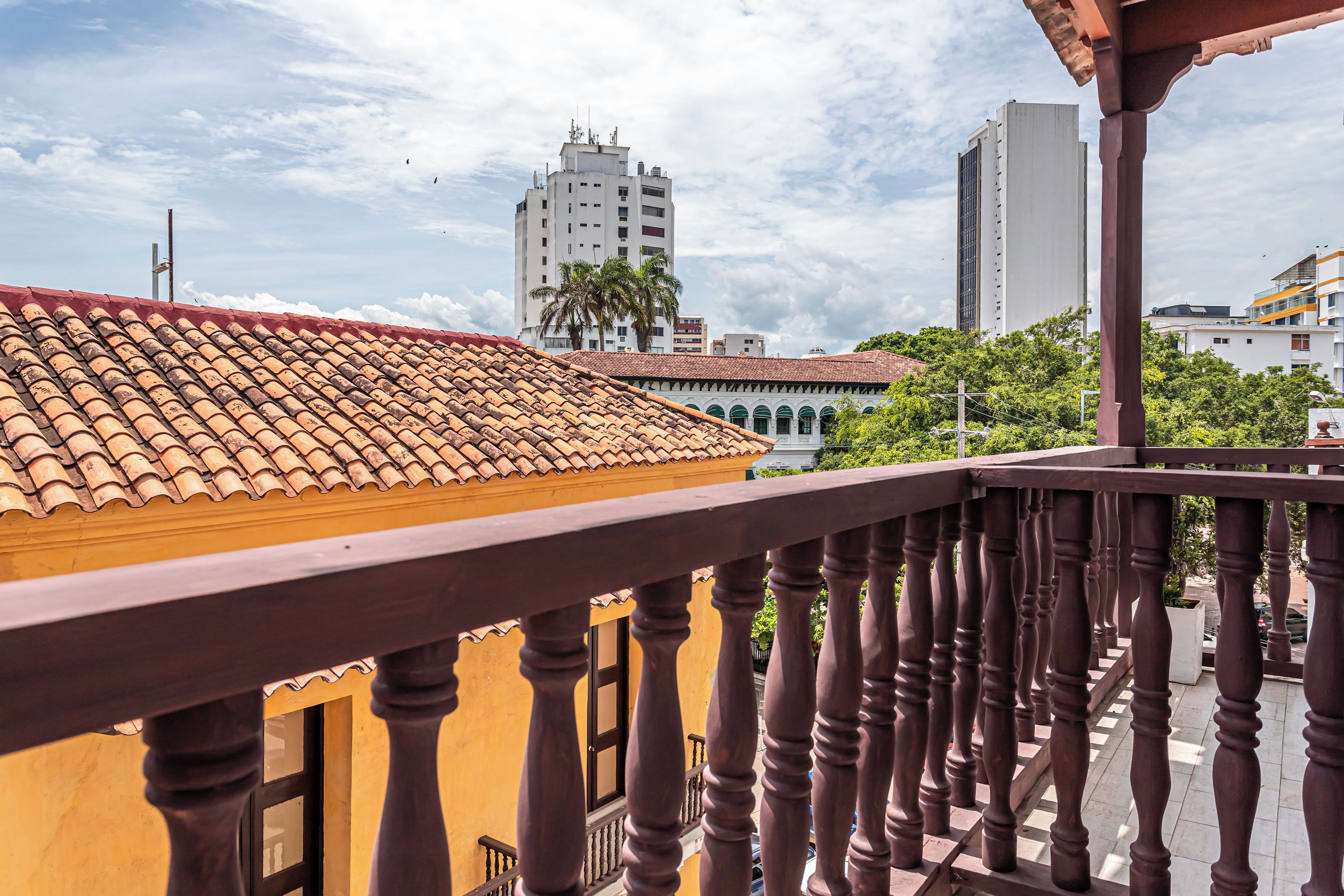 A balcony with a tiled roof, a wooden railing, and a view of a cityscape with buildings and palm trees.