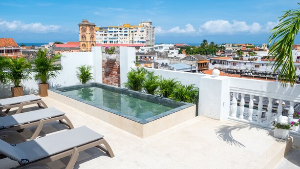 A rooftop pool area with lounge chairs and potted plants, overlooking a cityscape with buildings and a clear sky.