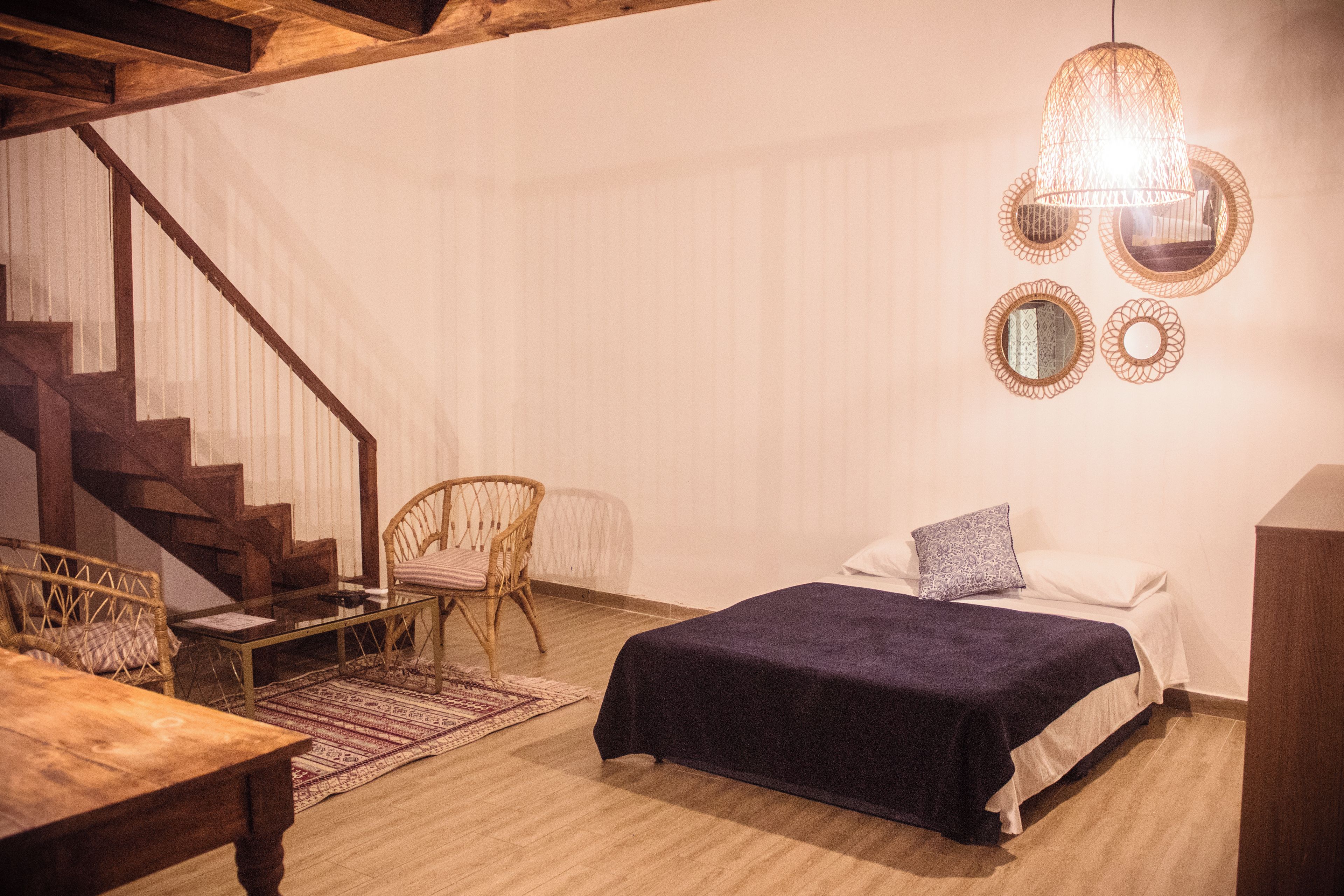 A room with a bed, a chair, a table, a staircase, and a hanging light fixture.
