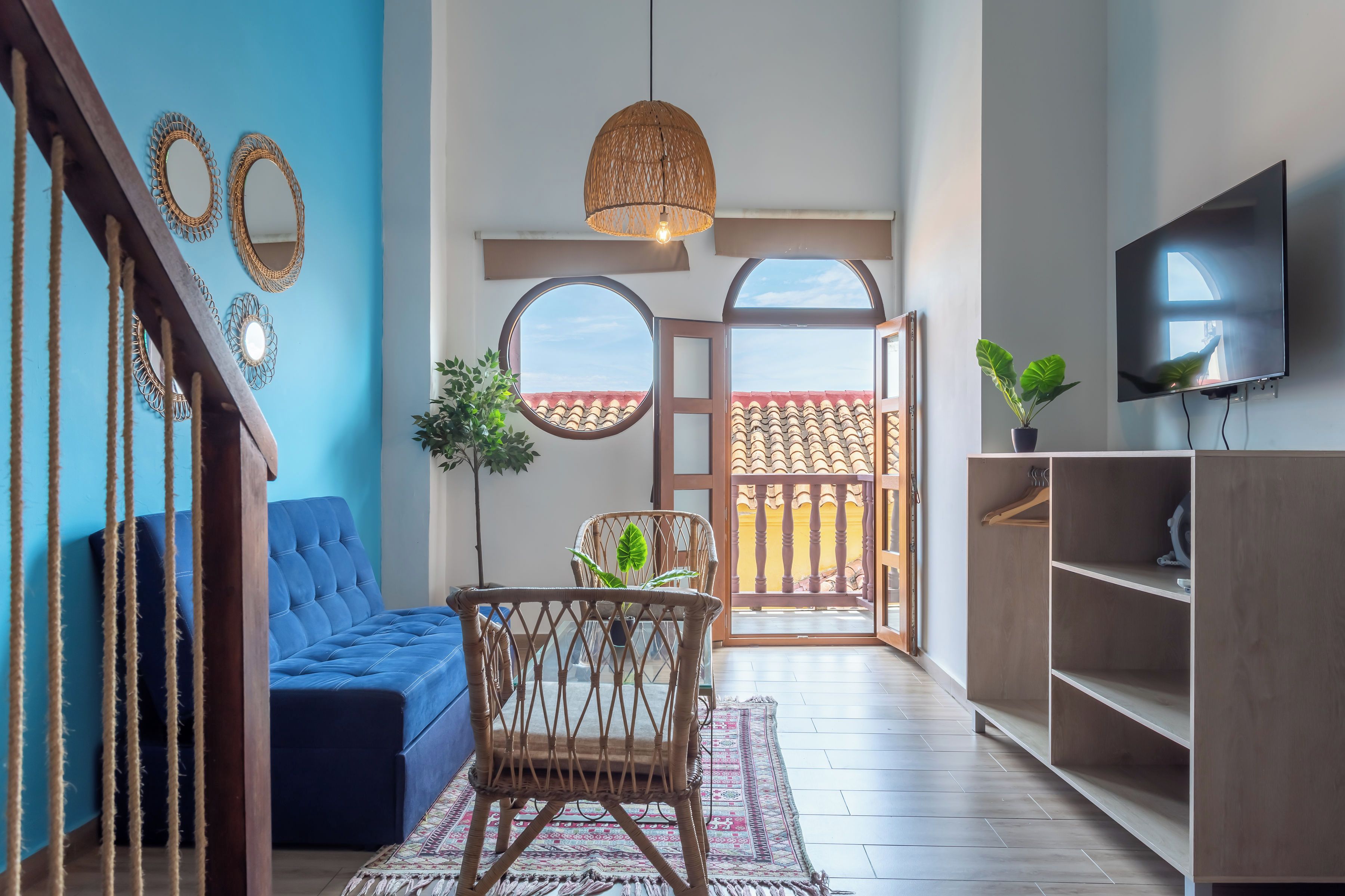 A modern living room with a blue sofa, a wicker chair, a round mirror, and a television on the wall.