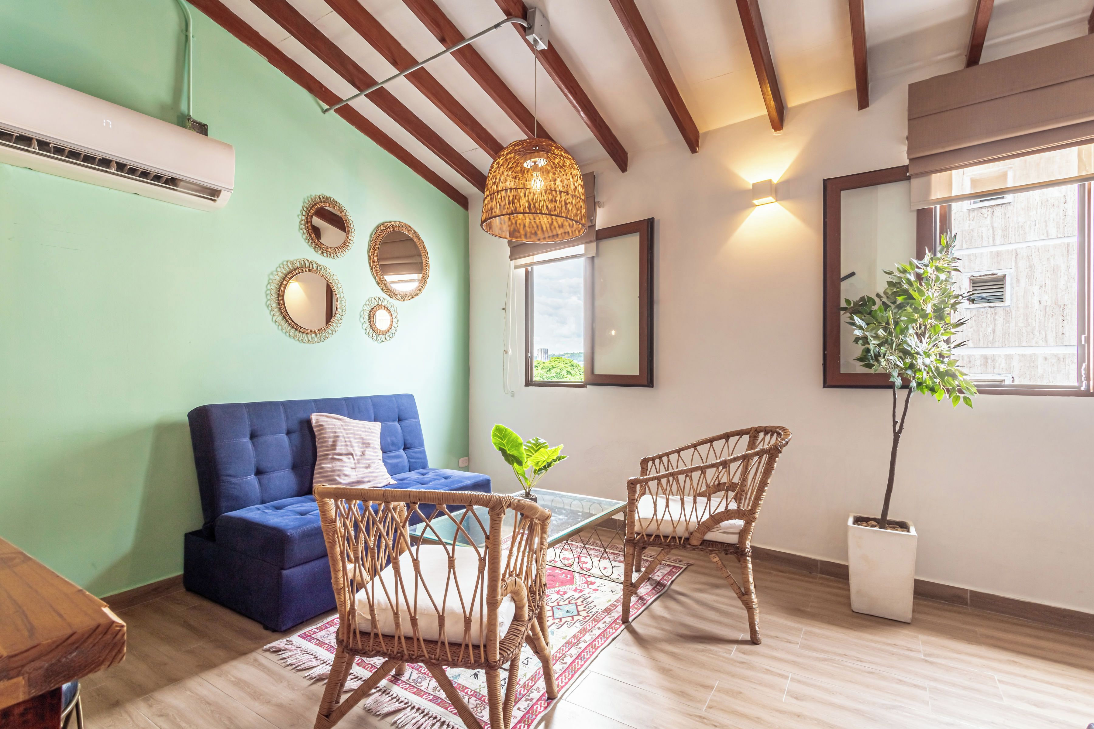 A room with a blue sofa, wicker chairs, a hanging light fixture, and a large window with a view.