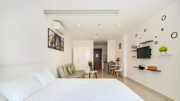 A modern hotel room with a large bed, a sofa, a small dining table, and a TV on the wall.