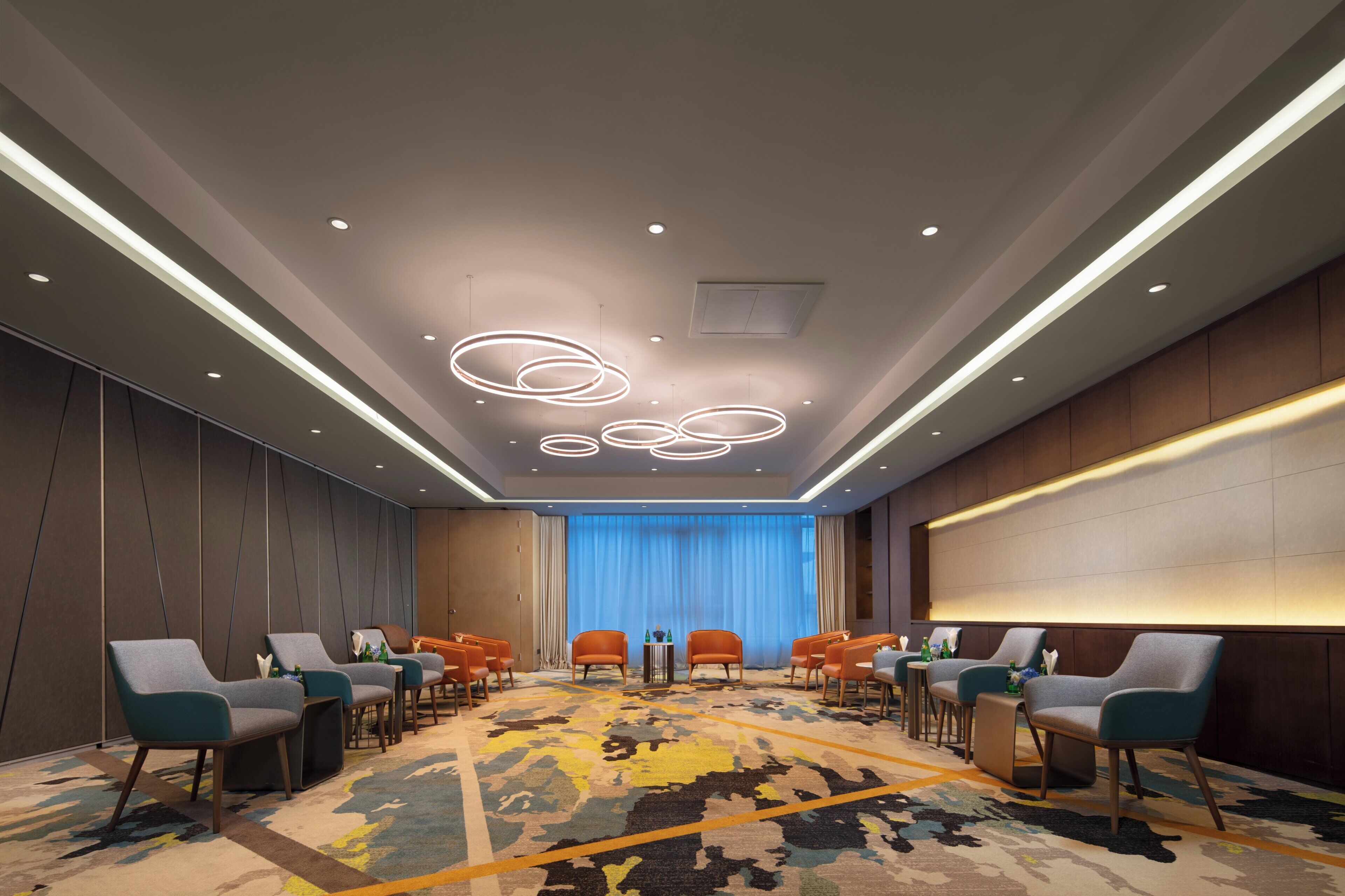 A conference room with a patterned carpet, multiple rows of chairs, a large screen, and a ceiling with circular light fixtures.