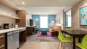 A modern kitchen with a dishwasher, a coffee machine, and a dining area with a table and chairs. - Home2 Suites by Hilton Silver Spring (Silver Spring)