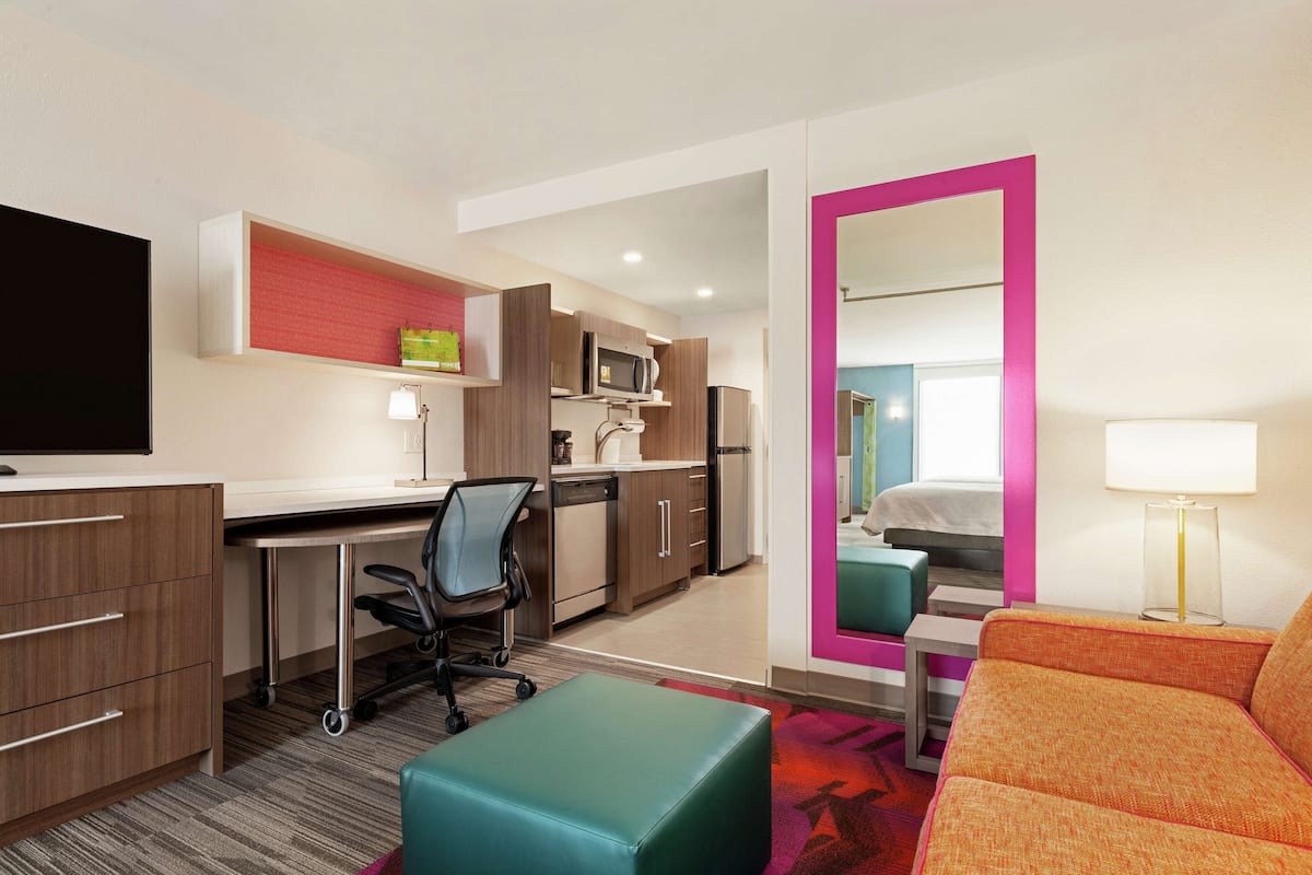 A hotel room with a kitchenette, a desk, a chair, a sofa, and a large mirror.