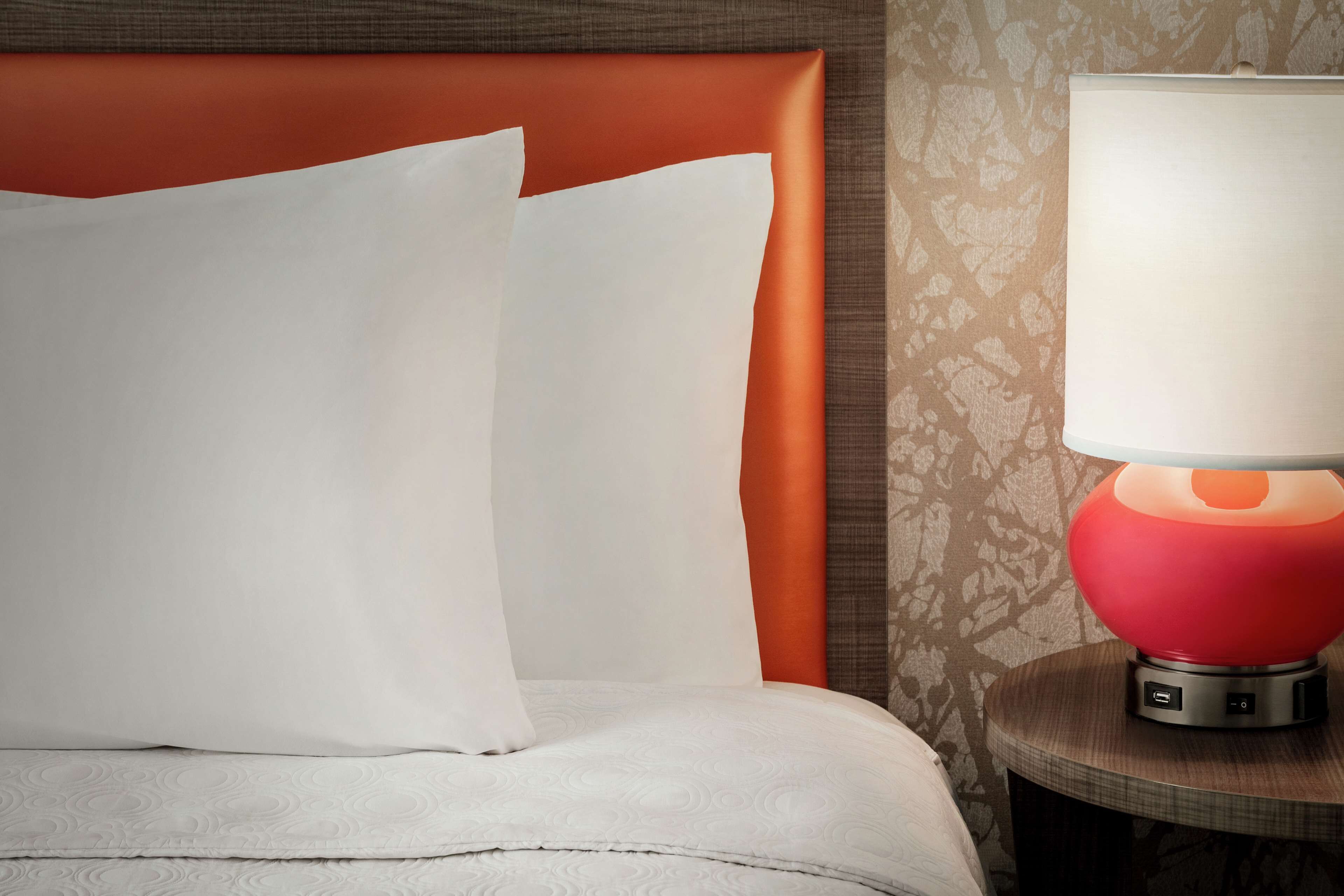 A neatly made bed with white linens and a red headboard, a round table with a red lamp, and patterned wallpaper.