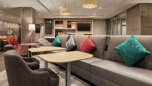 A modern lounge area with a sofa, chairs, and tables, featuring a flat-screen TV and a bookshelf. - Home2 Suites by Hilton Silver Spring (Silver Spring)