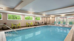 An indoor swimming pool with clear glass walls, green chairs, and tables. - Home2 Suites by Hilton Silver Spring (Silver Spring)