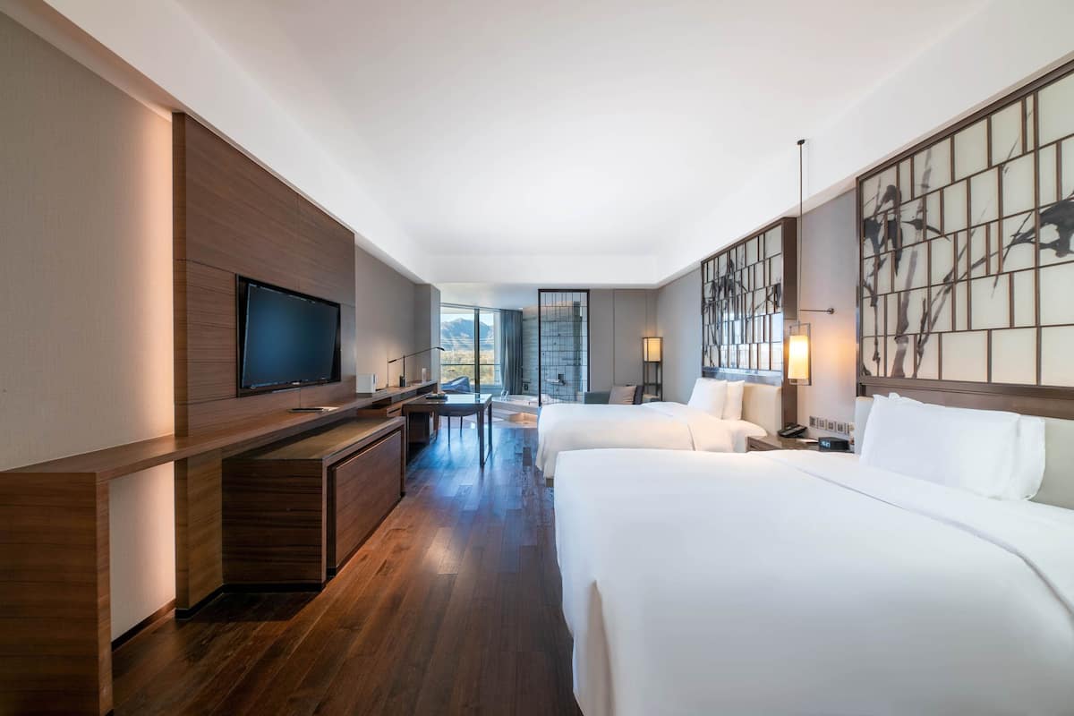 A hotel room with a large flat-screen TV, two beds, a desk, and a view of the mountains through large windows.