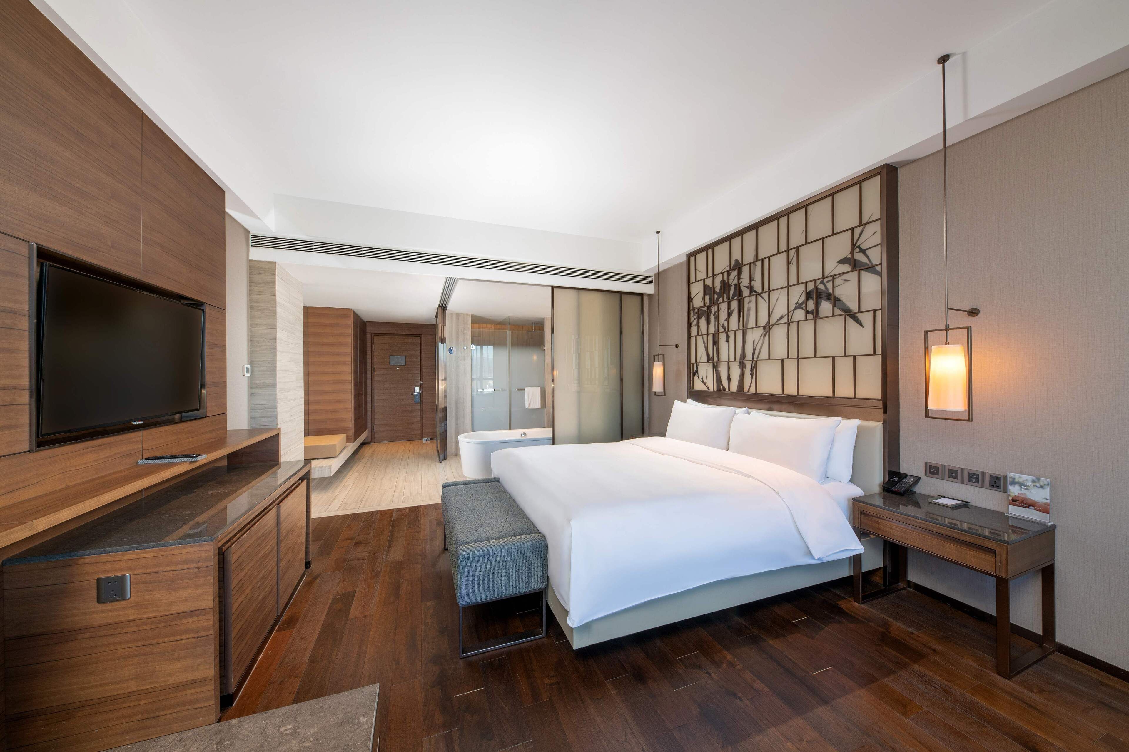 A modern hotel room with a large bed, a TV, a desk, and a bathroom visible through an open door.