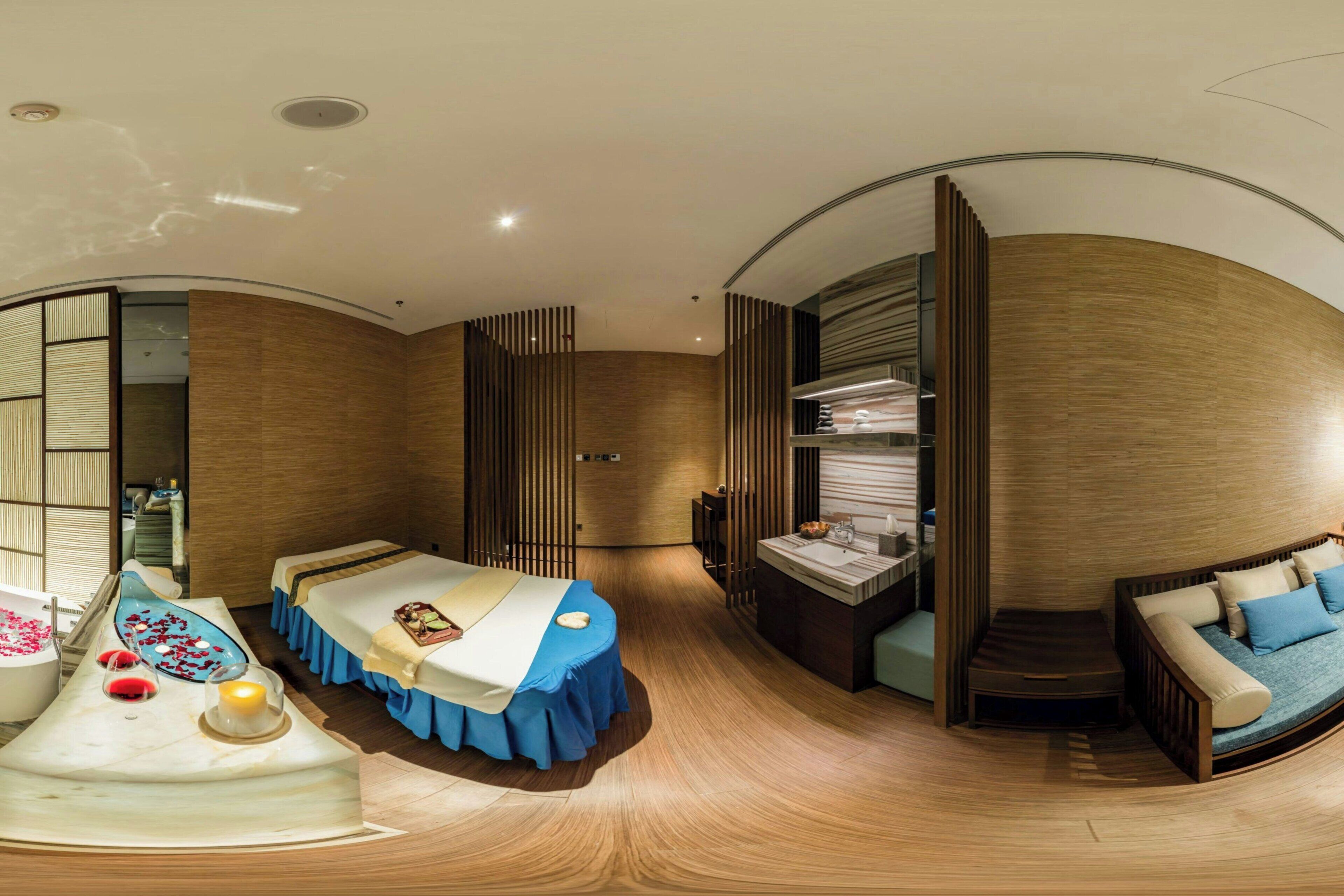 A modern hotel room with a curved layout, a bed with a blue bed skirt, a seating area with a sofa, a small table with a candle, and a built-in shelf with books.