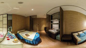 A modern hotel room with a curved layout, a bed with a blue bed skirt, a seating area with a sofa, a small table with a candle, and a built-in shelf with books.