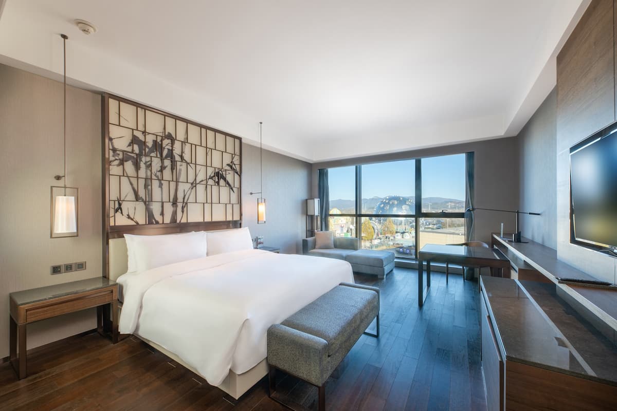 A modern hotel room with a large bed, a seating area, a TV, and a view of the city.