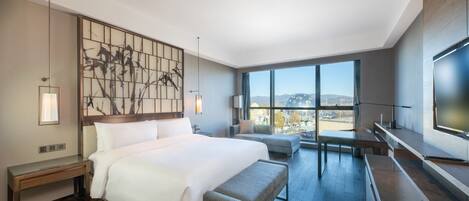 A modern hotel room with a large bed, a seating area, a TV, and a view of the city.