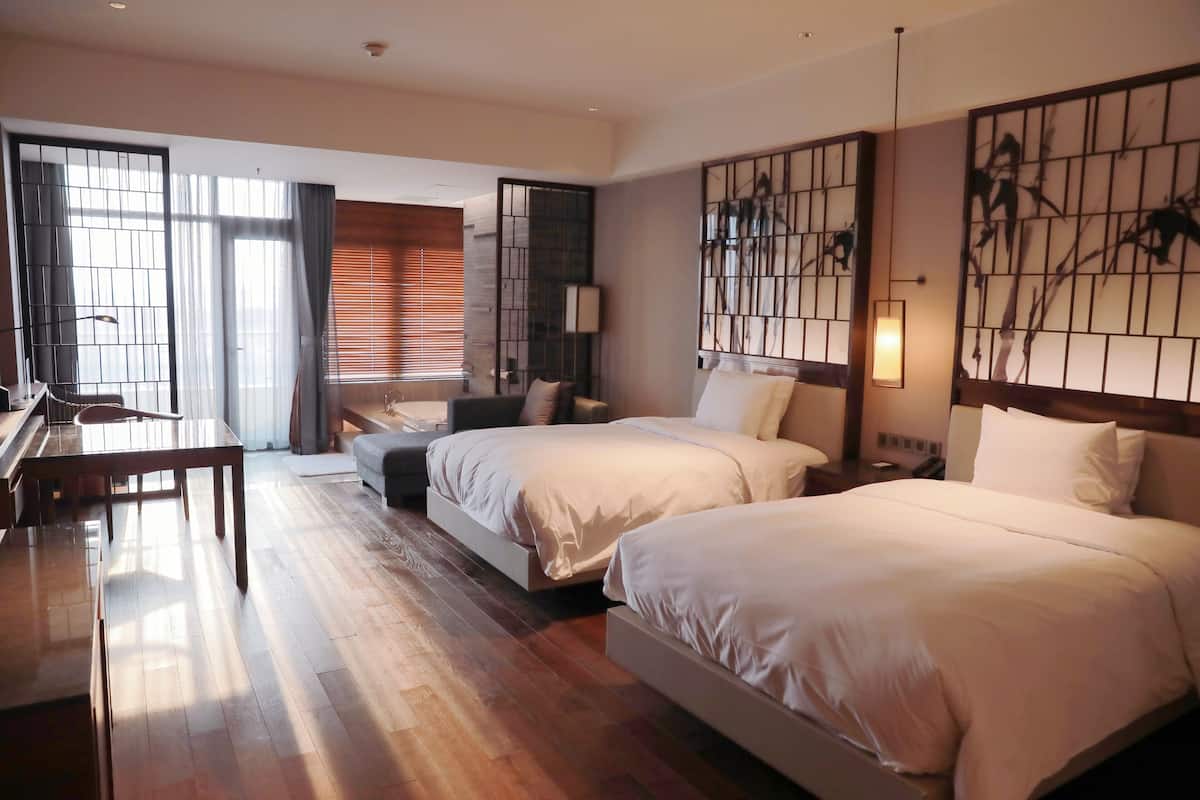 A hotel room with two beds, a desk, a chair, and a large window with curtains.