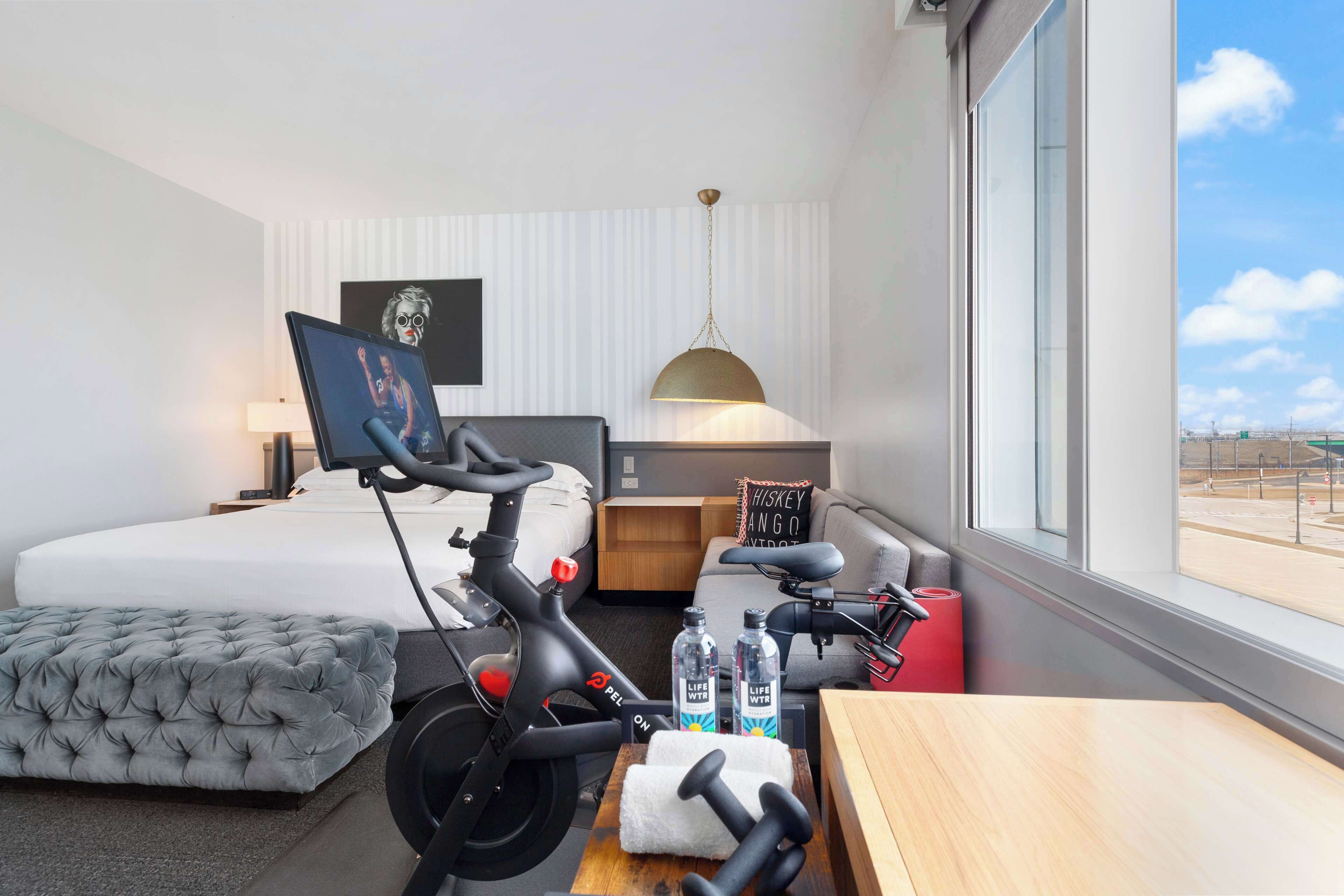 A hotel room with a bed, a grey tufted ottoman, a desk with dumbbells, a bicycle, and a view of the outside.