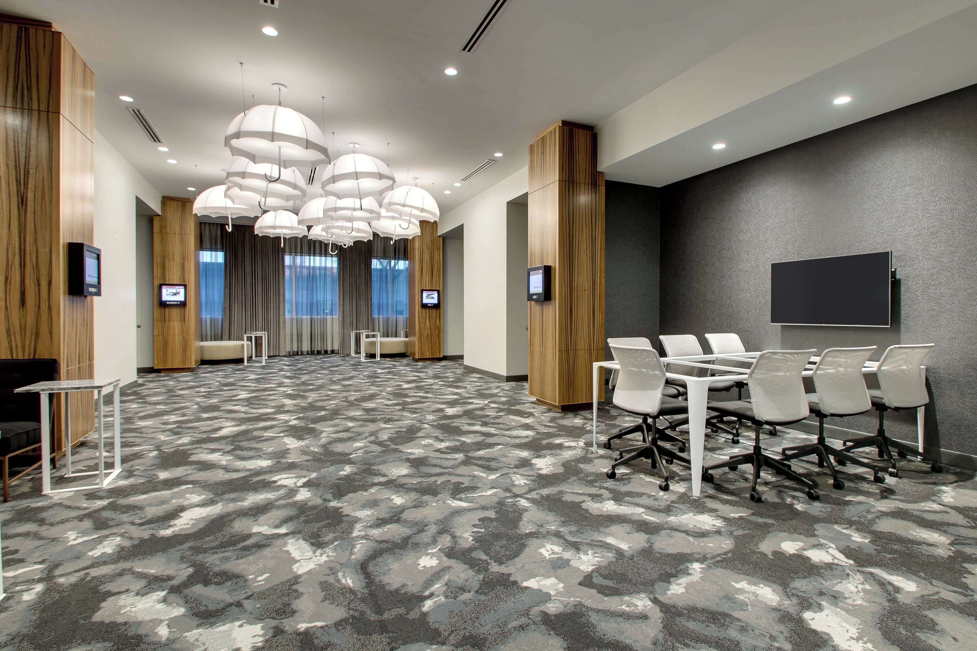 Une salle de conférence moderne avec un tapis à motifs gris, des chaises blanches, une grande table et de nombreux luminaires au plafond.
