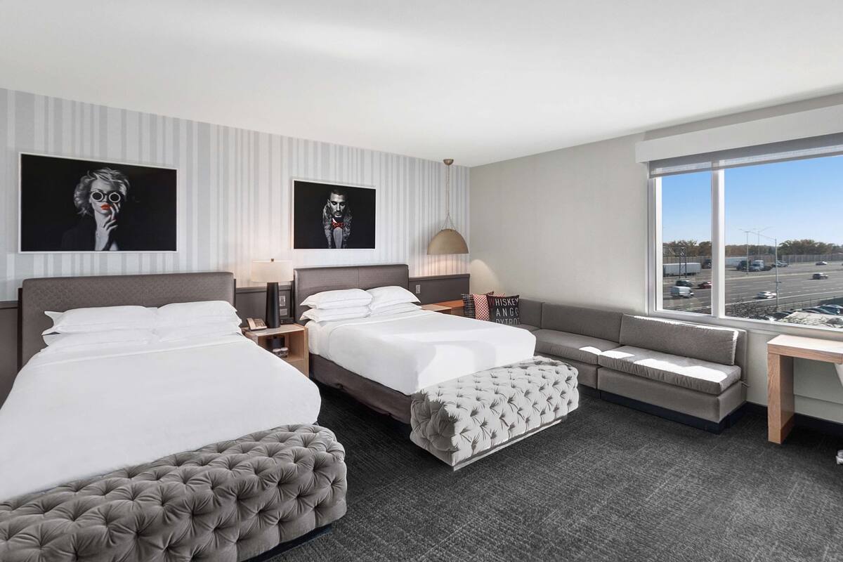 A modern hotel room with two beds, a sofa, a large window with a city view, and artwork on the walls.