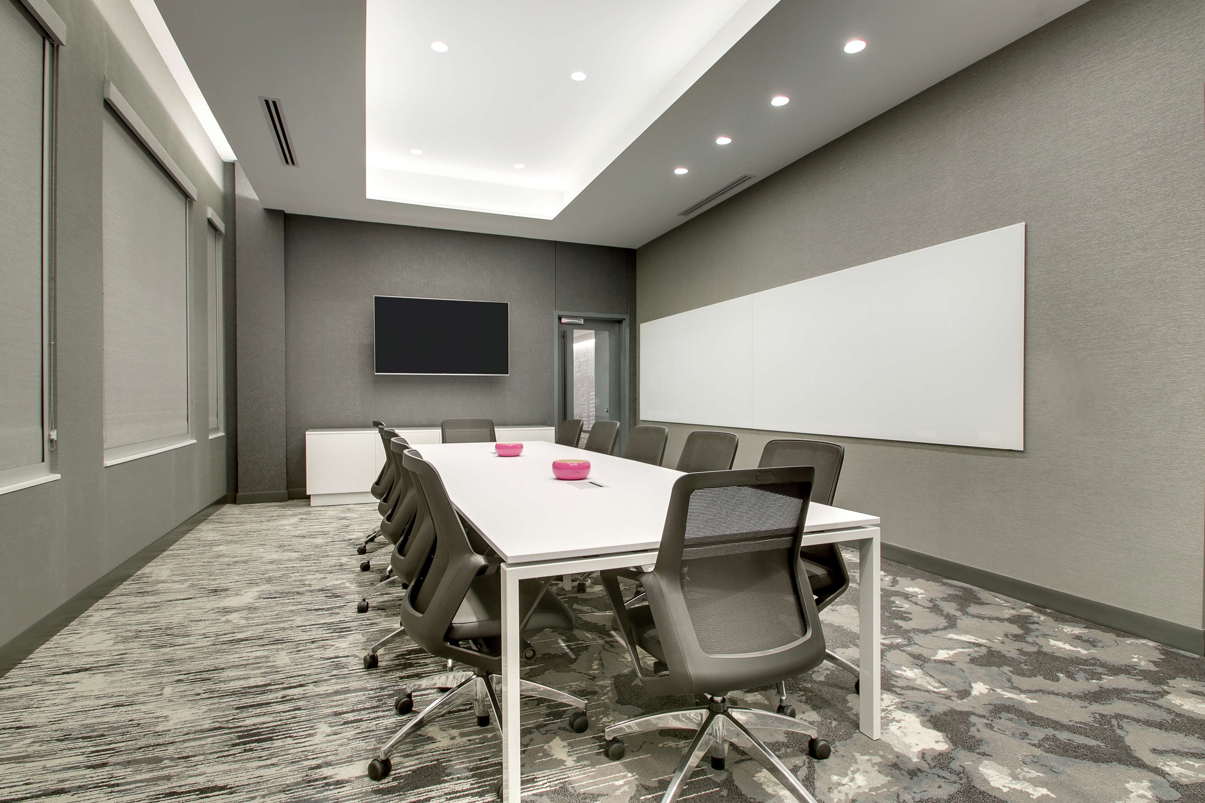 Un salón de conferencias con una mesa blanca, sillas grises, una pantalla grande y piso alfombrado.