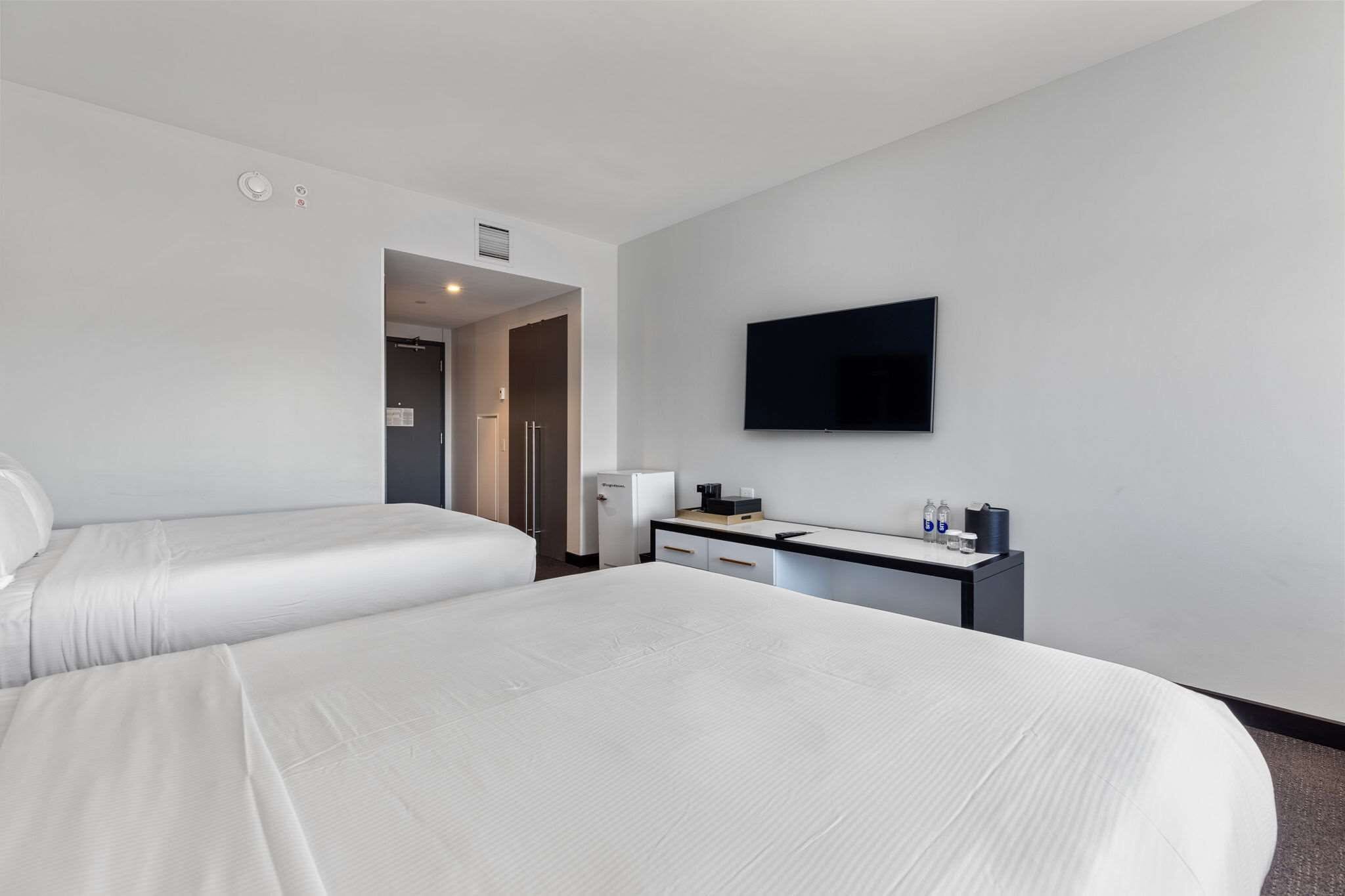 A hotel room with two beds, a desk, a flat-screen TV, and a mini-fridge.