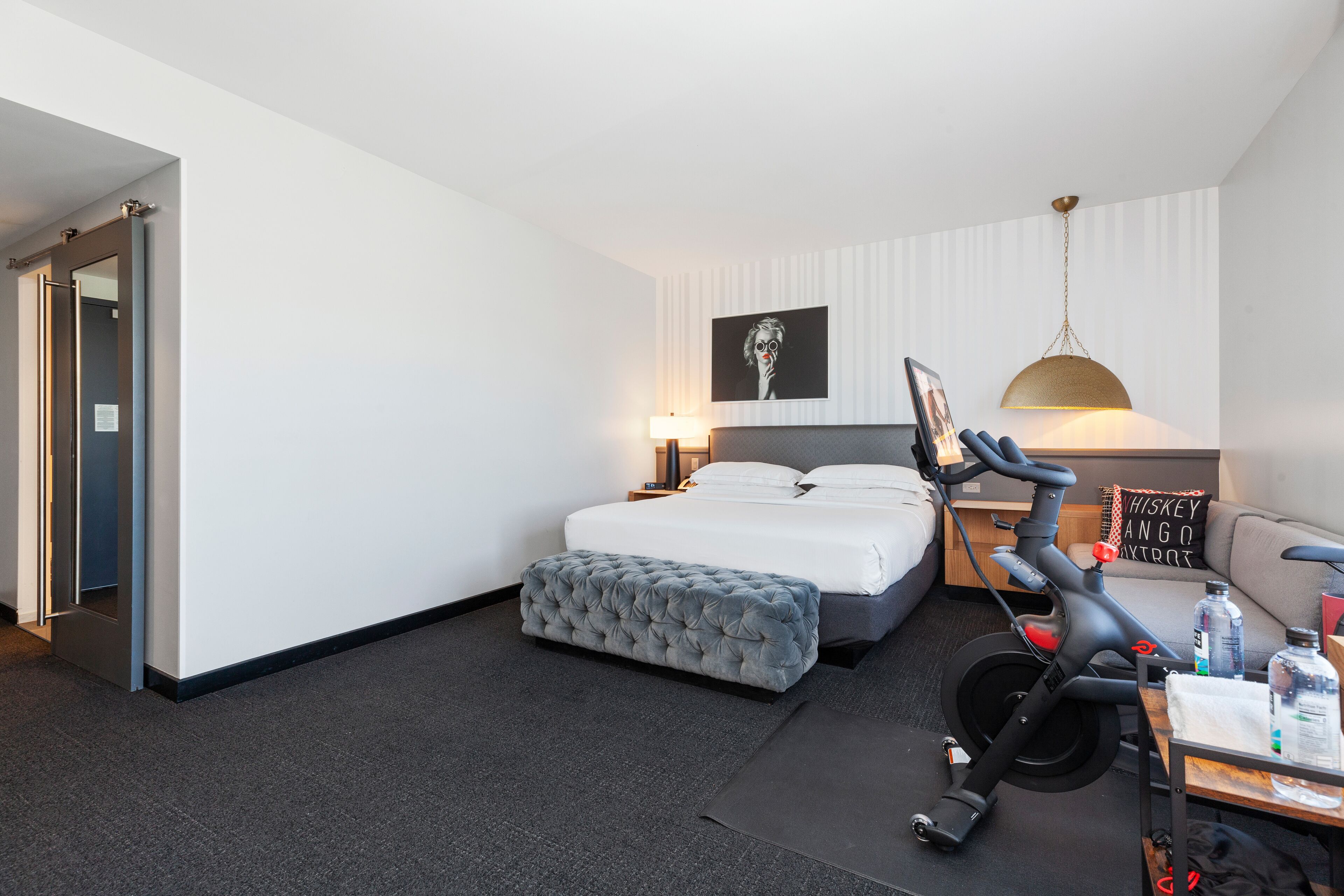 Quarto de hotel moderno com uma cama bem arrumada, um banquinho estofado cinza, uma escrivaninha com abajur e uma bicicleta fixada na parede.