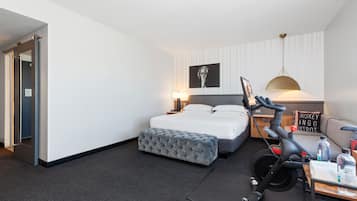 Quarto de hotel moderno com uma cama bem arrumada, um banquinho estofado cinza, uma escrivaninha com abajur e uma bicicleta fixada na parede.