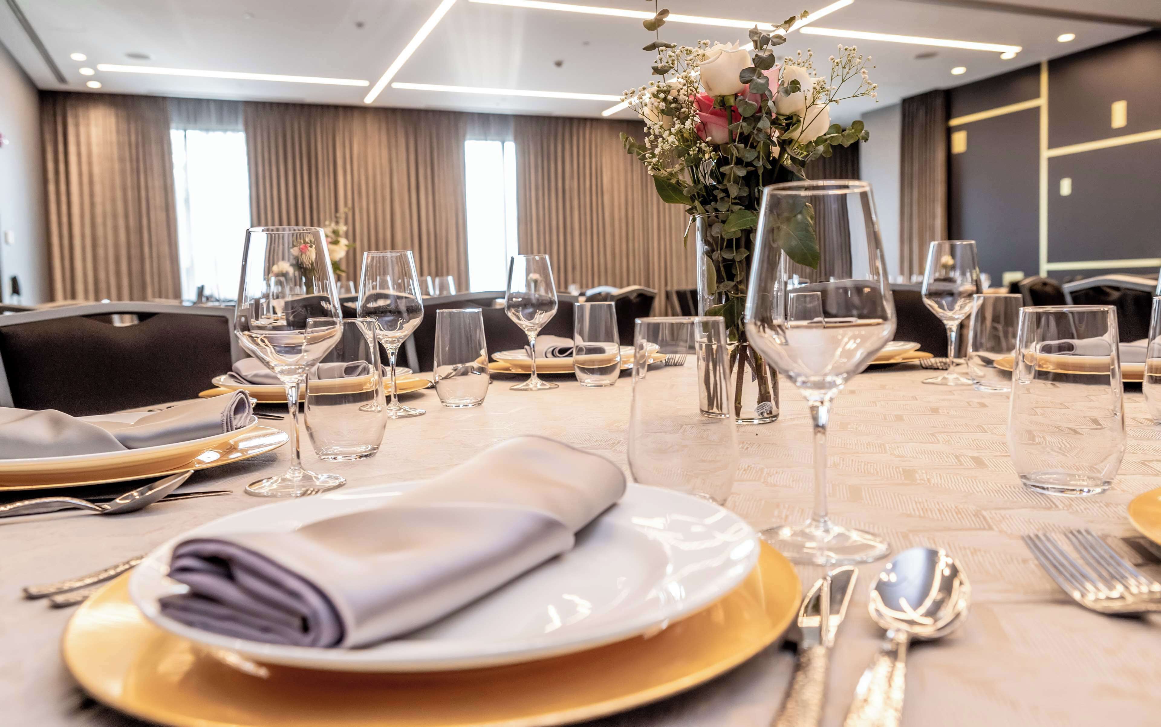 A hotel dining table set with white plates, gold chargers, and clear wine glasses, with a vase of flowers in the center.