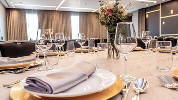 A hotel dining table set with white plates, gold chargers, and clear wine glasses, with a vase of flowers in the center.