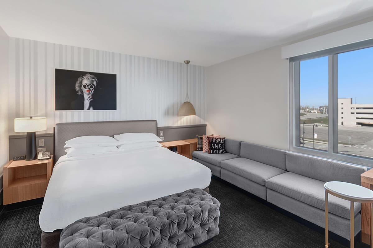 A hotel room with a large bed, a grey sofa, a small table, a lamp, and a view of the city through the window.