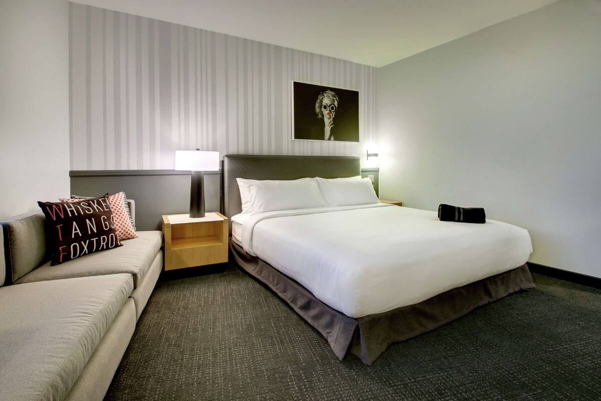A hotel room with a bed, a nightstand, a lamp, a pillow with text, and a wall with a striped pattern and a framed picture.