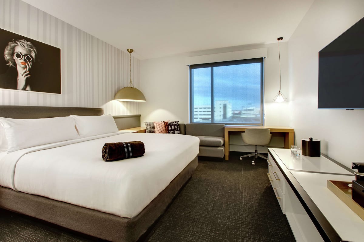 A modern hotel room with a large bed, a desk, a chair, a TV, and a window with a city view.