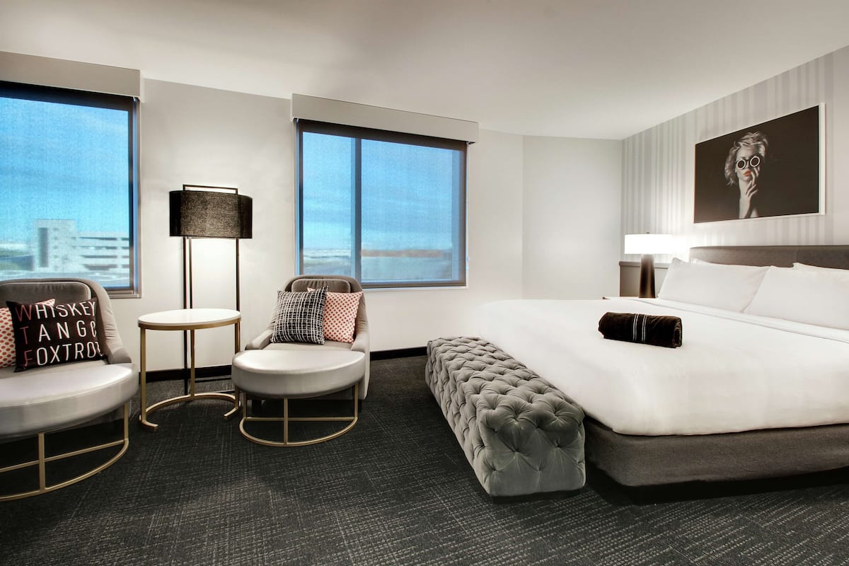 A hotel room with a large bed, a sofa, a small table, and a chair. There are two windows and a painting on the wall.