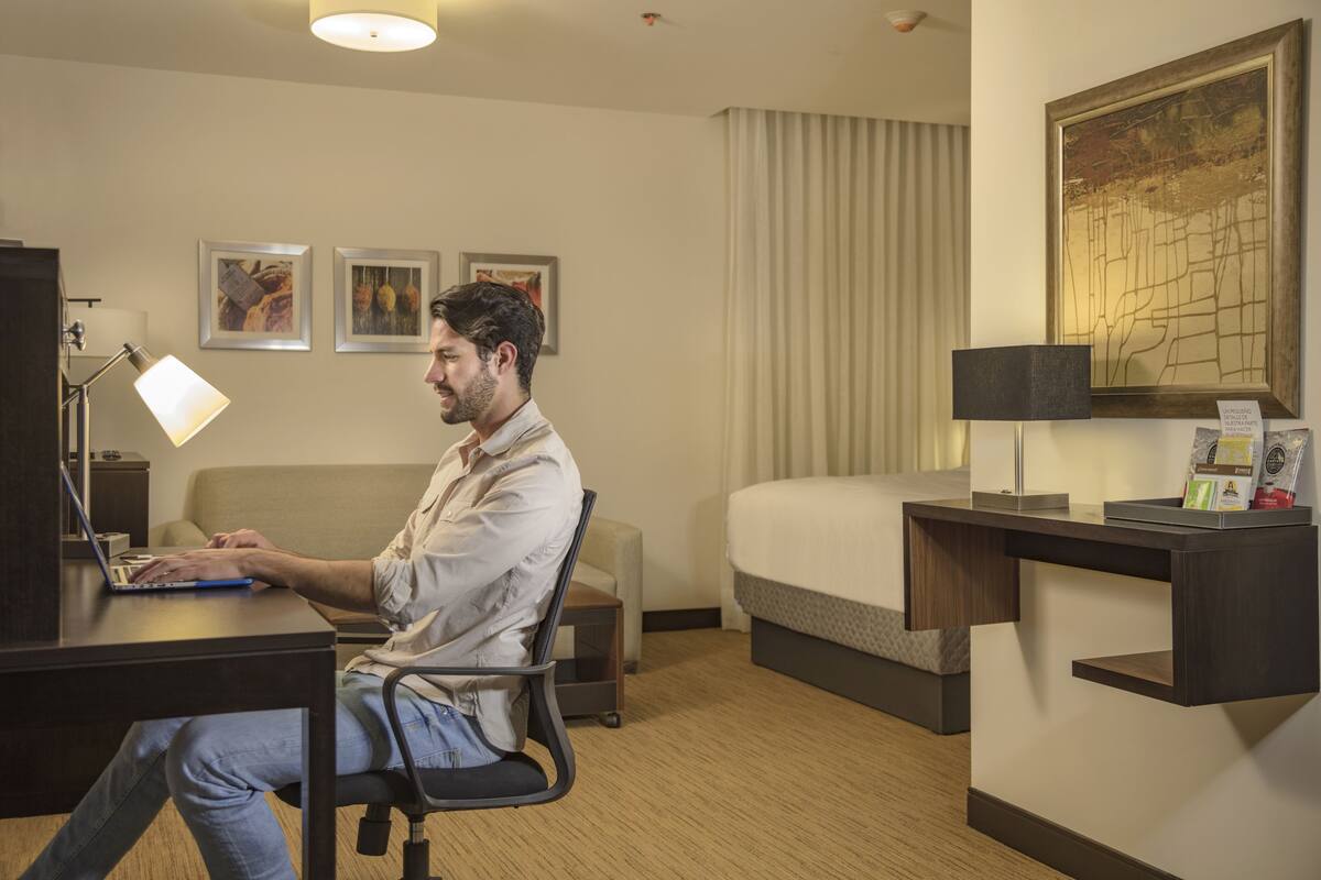 A hotel room with a desk, laptop, chair, bed, bedside table, lamp, and framed pictures on the wall.