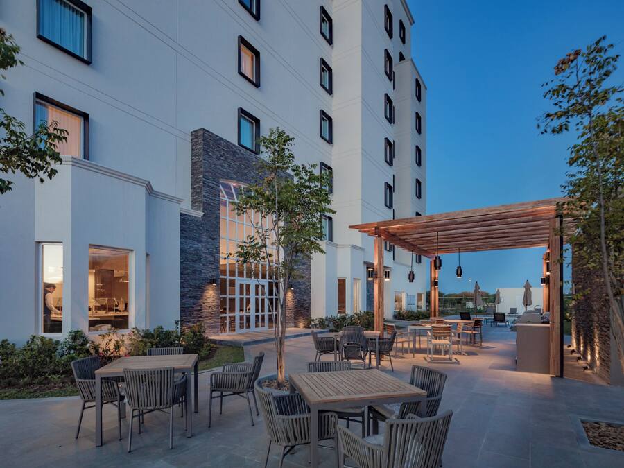 A modern outdoor patio area with multiple seating arrangements, a pergola, and a building with large windows and a stone facade.