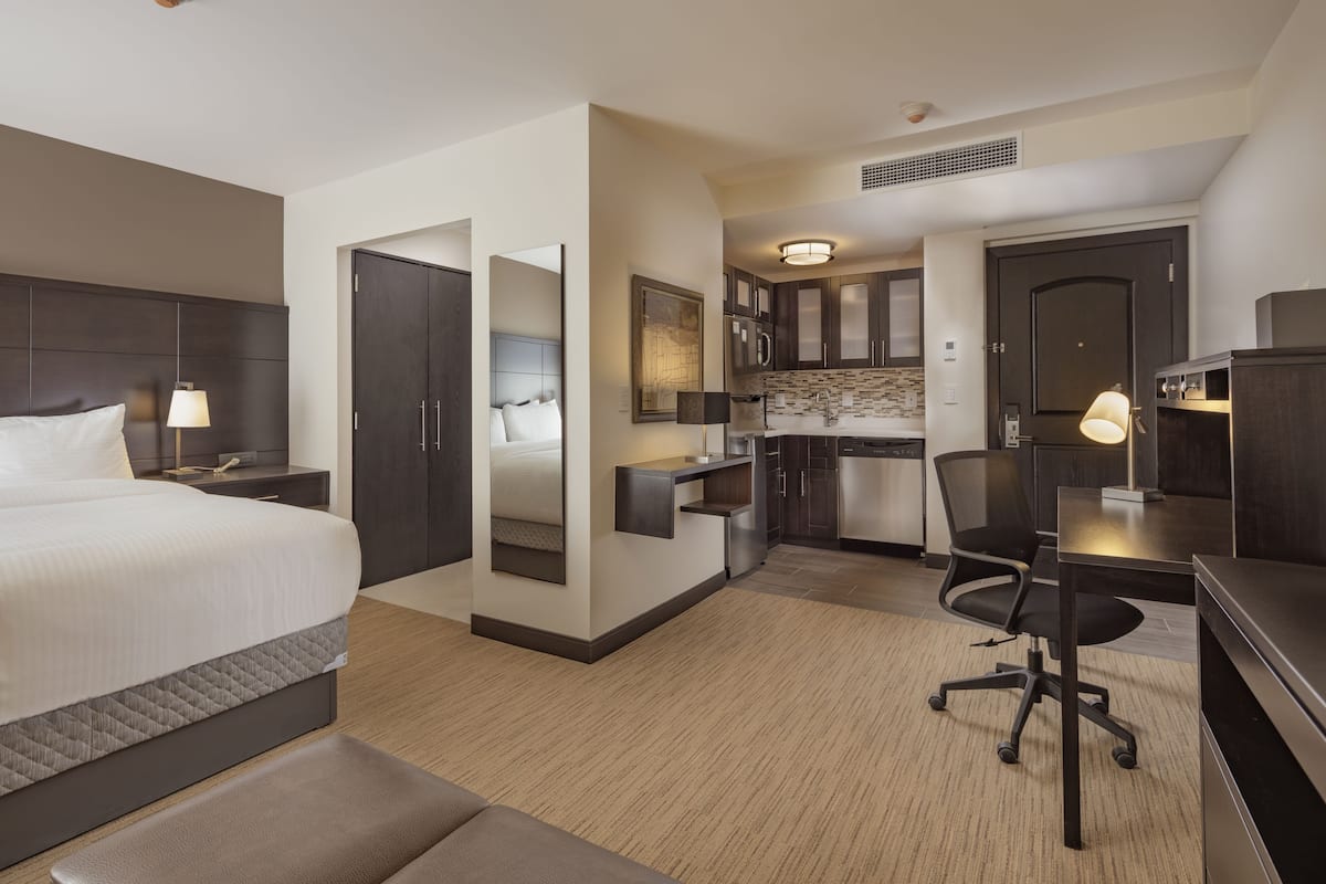 A hotel room with a bed, bedside table, lamp, desk, chair, kitchenette, and a door.