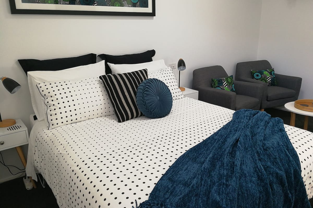 Un dormitorio ordenado con cama, una silla, una mesita y una lámpara de pared.