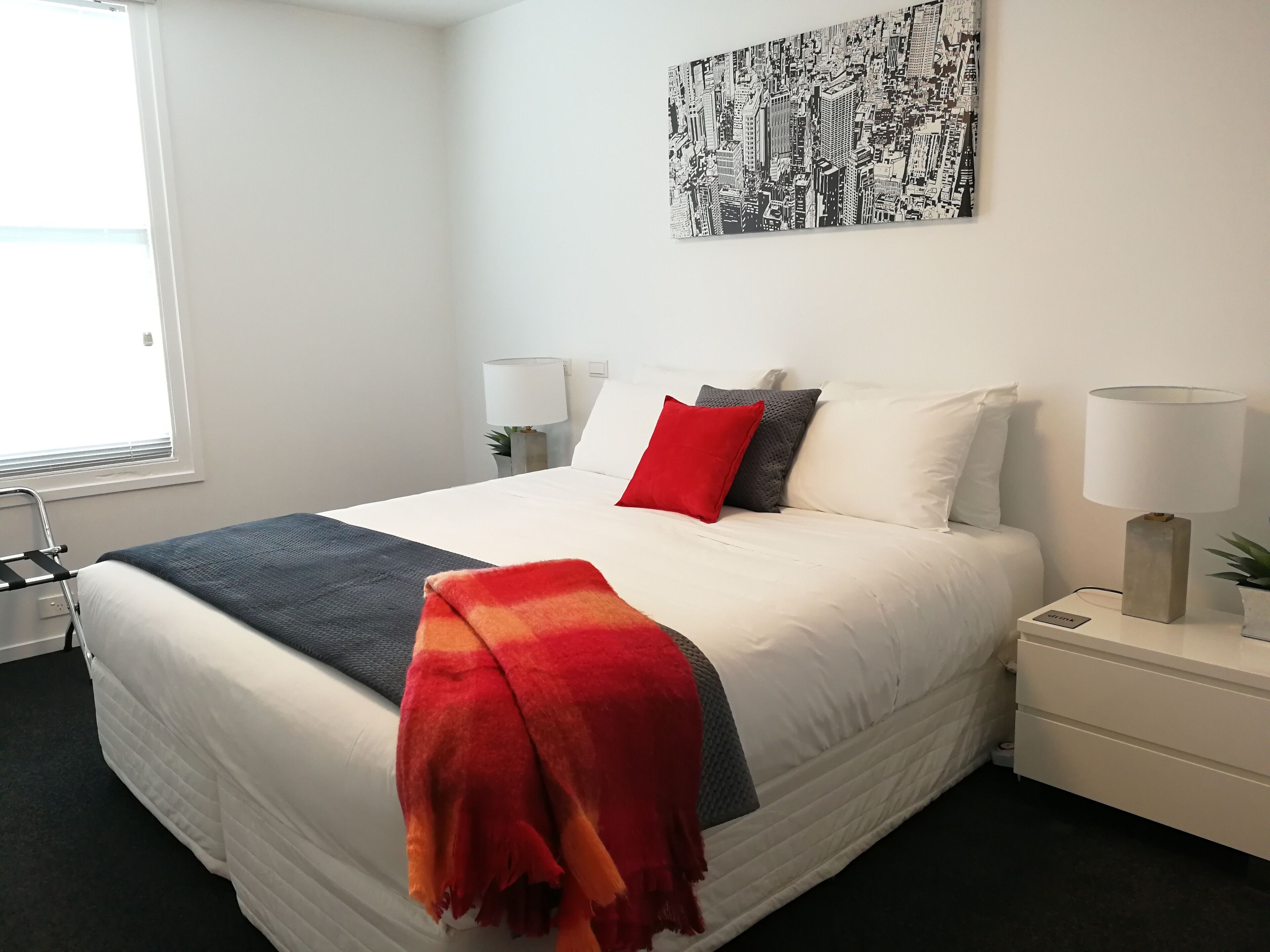 A neatly made bed with a white comforter and a red throw blanket, two bedside lamps, a small bedside table with a plant, and a cityscape artwork on the wall.